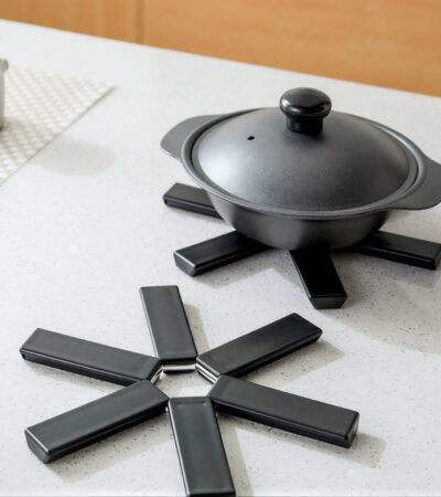 Black foldable heat insulation pot holder by Tawfeer showing the cross-shaped unfolded design on a kitchen counter.